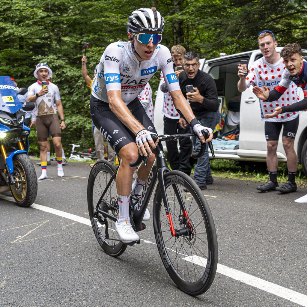 A cyclist in a white racing jersey and helmet rides a road bike on a tree-lined road, with spectators standing beside a van and a motorcycle nearby.