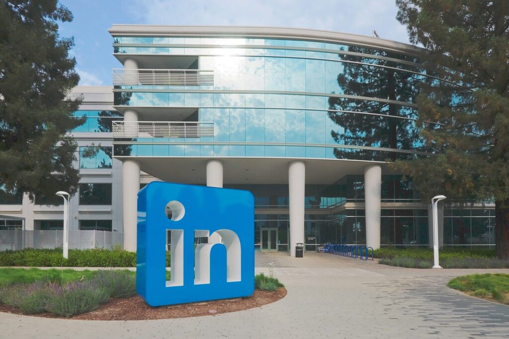 A large blue LinkedIn logo sign stands in front of a modern glass-and-white office building with trees, landscaping, and bicycle racks.