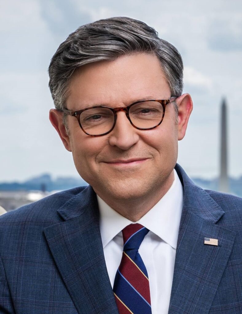 A close-up portrait of a man wearing glasses, a blue suit, striped tie, and U.S. flag lapel pin, with the Washington Monument visible in the blurred background.