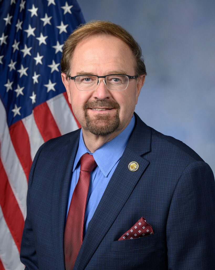 A formal portrait of a man wearing glasses, a dark suit jacket, blue shirt, and red tie, posed in front of a United States flag.