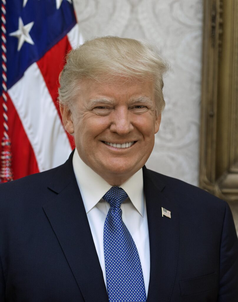 A formal portrait of Donald Trump wearing a dark suit and blue tie, with a U.S. flag behind him.
