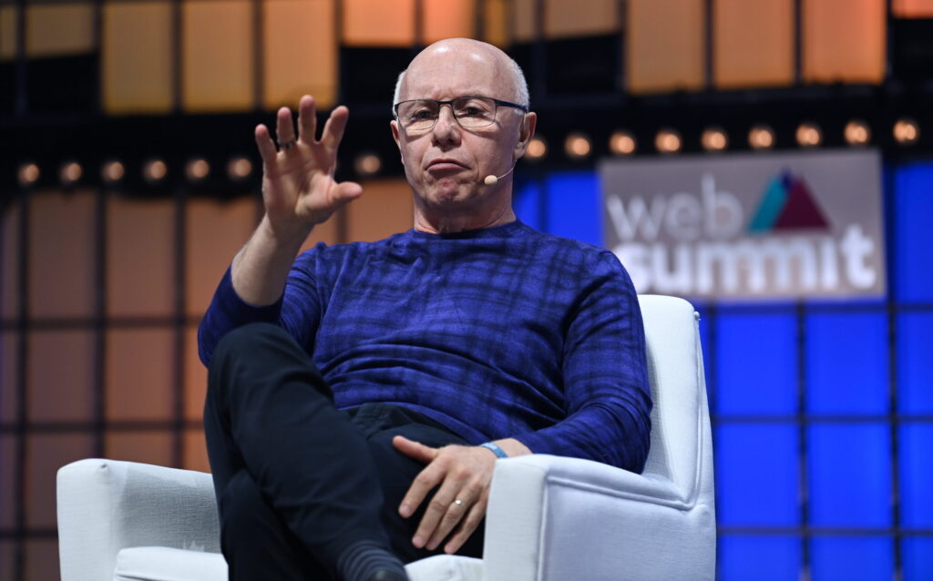 A bald man seated in a white armchair on a conference stage, raising one hand, with a Web Summit-style backdrop behind him.