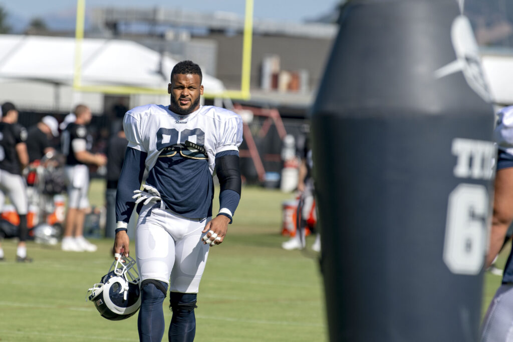 Aaron Donald in Rams practice gear walking on a field while carrying a football helmet; a large padded training object is in the foreground.
