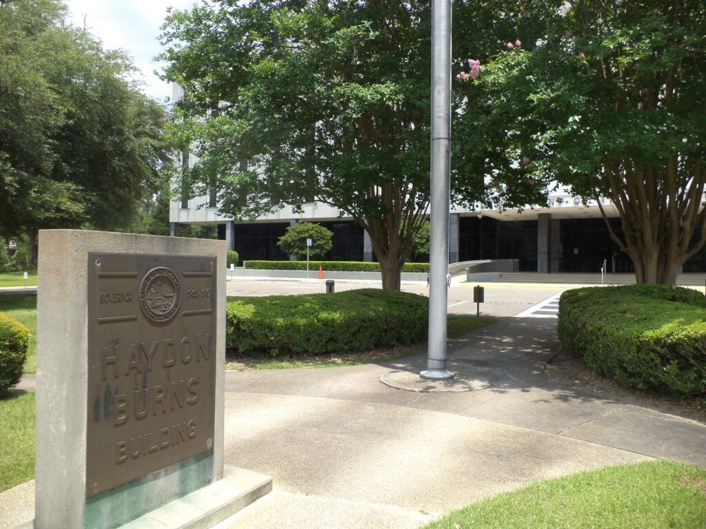 An exterior view of a Florida Department of Transportation building entrance area, with a freestanding agency sign in the foreground, trees and a flagpole.