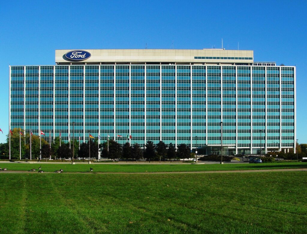 A wide exterior view of Ford's glass-fronted office building, with a Ford oval logo near the roofline and a large green lawn in front.