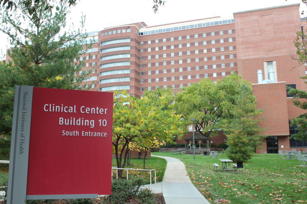 The entrance area of the NIH Clinical Center, including a red Clinical Center sign, landscaped walkway, and the large hospital building behind it.