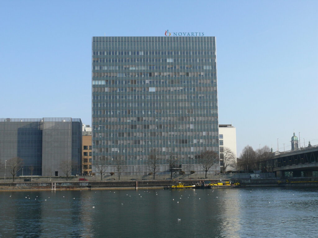 A modern glass-and-steel Novartis office building beside water, with smaller office buildings and trees nearby.