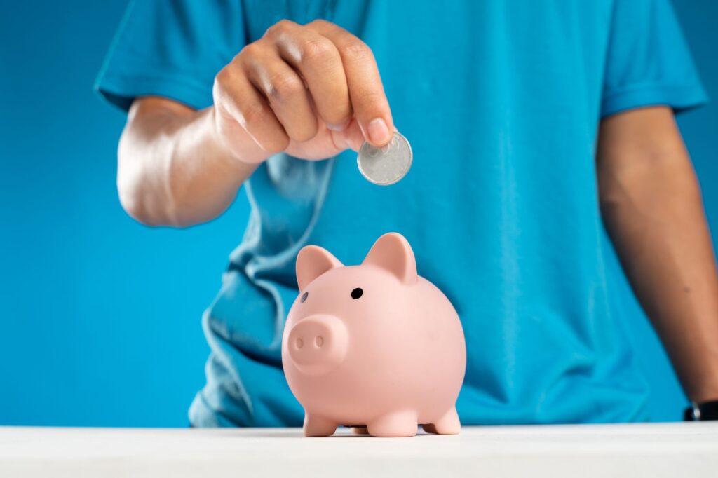 A close view of a person in a blue shirt placing a coin into a pink piggy bank.