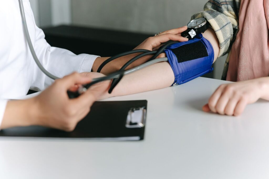 A healthcare worker measuring a seated patient's blood pressure with an arm cuff and manual sphygmomanometer on a desk in a clinical setting.