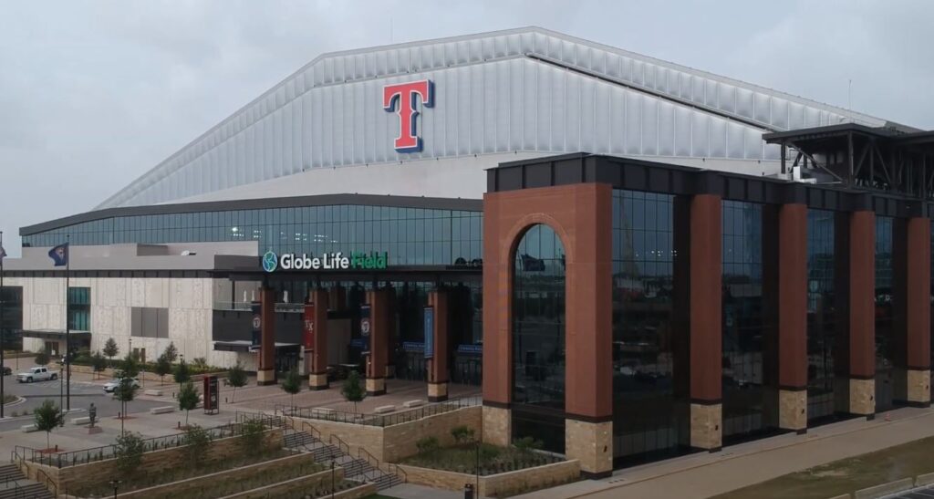 An exterior view of a large baseball stadium entrance, with a Texas Rangers-style red "T" logo high on the facade and brick columns in the foreground.