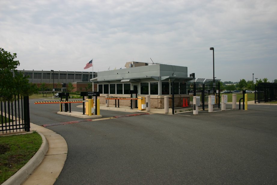 An exterior view of a low Federal Aviation Administration air-traffic-control facility, with a driveway, entrance barriers, fencing, flagpoles, and parked