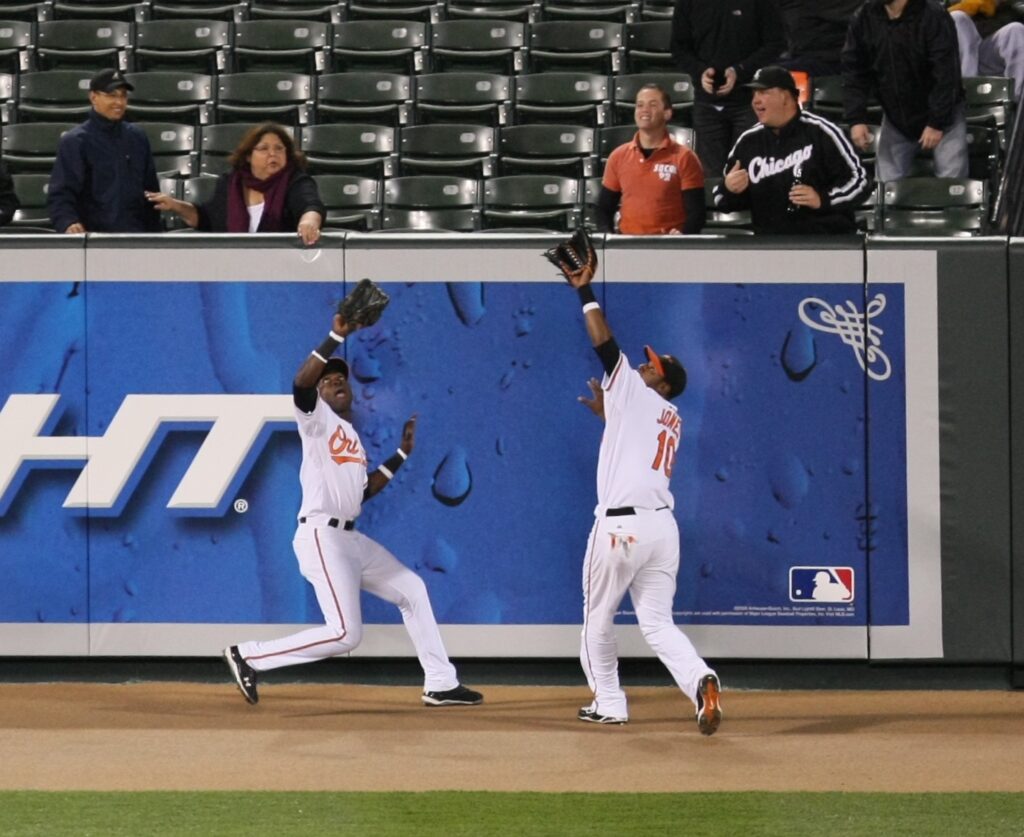 Two uniformed baseball outfielders colliding or converging near a padded outfield wall while attempting a play.
